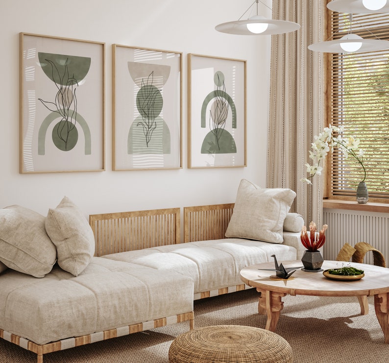 May include: A light beige sectional sofa with three cushions and a wooden frame. The sofa is in a living room with three framed abstract art prints with green and brown geometric shapes. A small round wooden coffee table with a black origami crane and a vase of red flowers sits in front of the sofa.