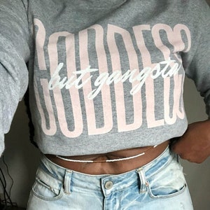 May include: A gray sweatshirt with the text "Goddess but gangsta" printed in pink. The sweatshirt is worn by a person wearing light blue jeans with a white beaded waist chain.