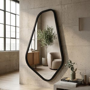 Può includere: Un grande specchio con cornice nera dalla forma irregolare è appeso a una parete chiara e strutturata. Lo specchio riflette una stanza con un mobile in legno, un albero di ulivo in vaso e una poltrona beige. Il testo "BLACK FRAME" è in basso.