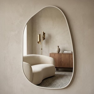 May include: An organically shaped mirror with a white frame is mounted on a neutral wall. The mirror reflects a room with a curved cream sofa, a wooden cabinet, and a gold floor lamp. The text "WHITE FRAME" is at the bottom.