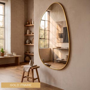 Può includere: Uno specchio di forma irregolare con cornice dorata è appeso a una parete di colore neutro. Lo specchio riflette una stanza con una finestra, mobili in legno e oggetti decorativi. Le parole "GOLD FRAME" sono in basso.