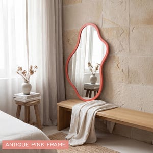 May include: An irregular, organic-shaped wall mirror featuring a smooth antique pink frame. The reflective glass surface captures a view of a room, and the text "ANTIQUE PINK FRAME" is printed in white on a pink rectangle at the bottom.