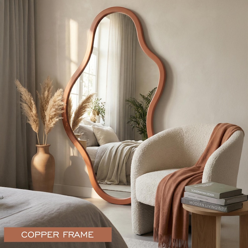 May include: A large, floor-standing mirror featuring an irregular, organic, wavy shape and a smooth copper-colored frame. The mirror reflects a bedroom interior, including a bed with neutral bedding and a green plant, while standing on the floor.