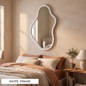 May include: A wall-mounted mirror featuring an irregular, organic, wavy shape and a smooth white frame. The reflective glass surface captures the surrounding room, and the text "WHITE FRAME" is clearly visible at the bottom of the image.