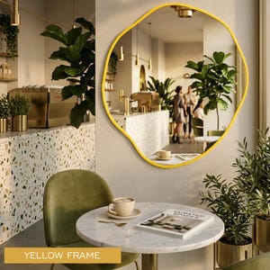 Peut inclure: Un miroir mural dot&eacute; d'une forme organique irr&eacute;guli&egrave;re et d'un cadre jaune vif. La surface r&eacute;fl&eacute;chissante offre une vue claire sur l'int&eacute;rieur d'un caf&eacute;, incluant une table en marbre, une chaise verte et des plantes en pot.