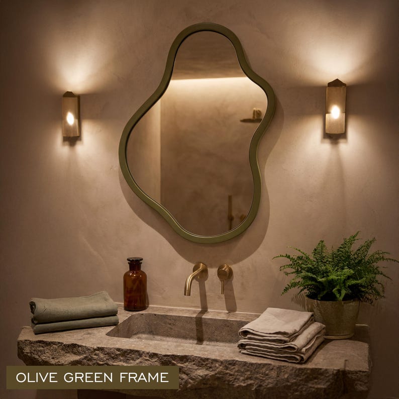 May include: An irregular, organic-shaped wall mirror featuring a smooth olive green frame. The mirror is mounted above a stone sink basin with a brass faucet, flanked by two wall-mounted light fixtures that cast a warm glow on the wall.