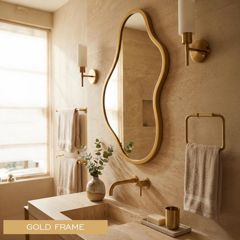 May include: An irregular, organic-shaped wall mirror featuring a smooth gold frame. The mirror is mounted on a light beige stone wall above a matching stone vanity, reflecting the surrounding bathroom space with its clear, reflective glass surface.