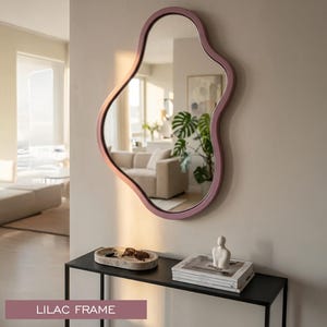 May include: An irregular, organic-shaped wall mirror featuring a smooth lilac frame. The mirror reflects a bright interior space with a sofa and a potted plant. The text LILAC FRAME is displayed at the bottom of the image.