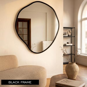 May include: A large, irregularly shaped mirror with a black frame hangs on a cream-coloured wall. The mirror reflects a room with a wooden door, a black metal shelf, and a beige armchair. The text "BLACK FRAME" is at the bottom of the image.