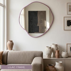 Peut inclure: Un miroir mural de forme organique et irr&eacute;guli&egrave;re dot&eacute; d'un cadre lisse de couleur lilas. La surface r&eacute;fl&eacute;chissante offre une vue claire de la pi&egrave;ce, incluant une peinture abstraite sombre sur le mur oppos&eacute;, capturant la lumi&egrave;re et la profondeur.