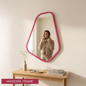 May include: A wall-mounted mirror featuring an irregular, organic-shaped frame in a vibrant magenta color. The reflective surface shows a reflection of a person wearing a beige sweater. The mirror is positioned above a wooden console table.