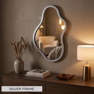 May include: An irregular, organic-shaped wall mirror featuring a smooth silver frame. The reflective surface displays a room interior with a chair and lighting. The text "SILVER FRAME" is printed in the lower left corner of the image.