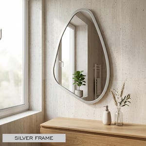 May include: An irregularly shaped mirror with a silver frame hangs on a textured wall. The mirror reflects a window and a small potted plant. A wooden cabinet below holds a soap dispenser and a vase with decorative stems. Text on the bottom reads "SILVER FRAME."