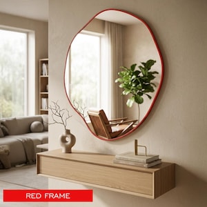 May include: A wall-mounted mirror featuring an irregular, organic-shaped frame in a vibrant red color. The reflective surface displays a clear view of a room interior, including a wooden chair and a green plant, with a red frame label at the bottom.