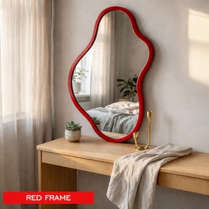 May include: A wall mirror with an irregular, organic-shaped red frame. The mirror reflects a bedroom interior with a bed and a plant. The text RED FRAME is visible at the bottom of the image, indicating the color of the mirror border.