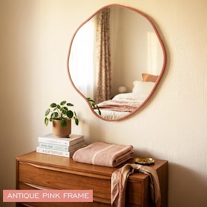Peut inclure: Un miroir mural de forme organique irr&eacute;guli&egrave;re dot&eacute; d'un cadre rose antique. Le miroir refl&egrave;te l'int&eacute;rieur d'une chambre, incluant un lit avec des oreillers et un rideau. Le cadre pr&eacute;sente un bord lisse et continu avec une silhouette arrondie.