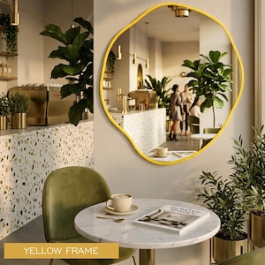 Peut inclure: Un miroir mural doté d'un cadre aux formes organiques et irrégulières avec une finition jaune vif. La surface réfléchissante offre une vue claire sur l'intérieur d'un café, incluant un comptoir en terrazzo, des plantes et du mobilier.