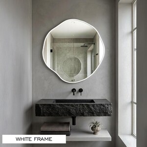 Peut inclure: Un miroir mural de forme organique irr&eacute;guli&egrave;re dot&eacute; d'un cadre blanc fin. La surface r&eacute;fl&eacute;chissante montre l'int&eacute;rieur d'une salle de bain avec un lavabo en pierre textur&eacute;e sombre, des robinets muraux noirs et une petite plante en pot.