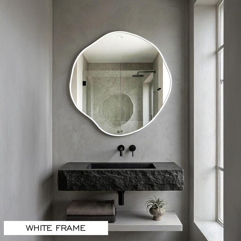 Peut inclure: Un miroir mural de forme organique irr&eacute;guli&egrave;re dot&eacute; d'un cadre blanc fin. La surface r&eacute;fl&eacute;chissante montre l'int&eacute;rieur d'une salle de bain avec un lavabo en pierre textur&eacute;e sombre, des robinets muraux noirs et une petite plante en pot.
