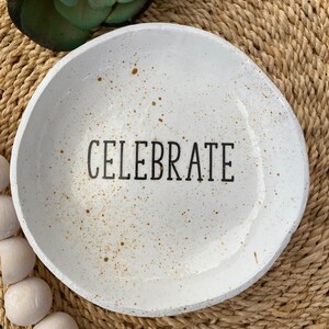 Puede incluir: Bol de cer&aacute;mica blanco con la palabra "CELEBRATE" en letras negras. El cuenco tiene un dise&ntilde;o moteado con detalles en marr&oacute;n. El cuenco es redondo y tiene un borde ligeramente irregular. El cuenco est&aacute; sobre una superficie tejida.
