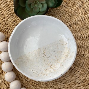 Puede incluir: Un cuenco de cer&aacute;mica blanca con un patr&oacute;n dorado moteado en la mitad inferior. El cuenco tiene un borde rugoso y texturizado y est&aacute; sobre una estera tejida. Una suculenta verde es visible en el fondo.