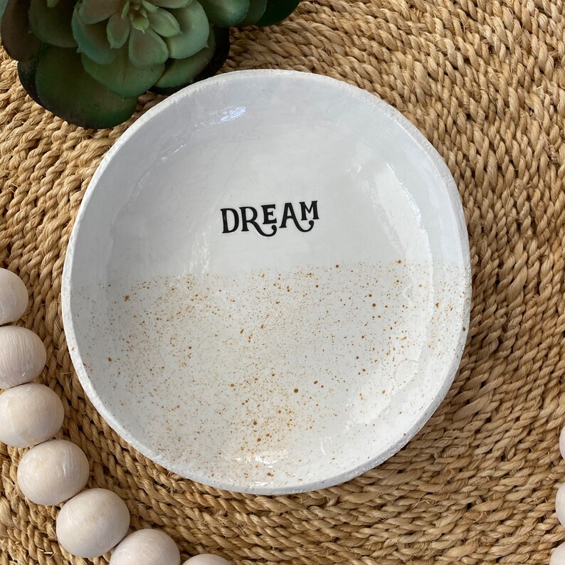 Puede incluir: Un cuenco de cer&aacute;mica blanco con la palabra "DREAM" en escritura negra. El cuenco tiene un dise&ntilde;o moteado y texturizado con motas marrones. El cuenco es redondo y tiene un borde ligeramente elevado. El fondo es una estera de fibra natural tejida.