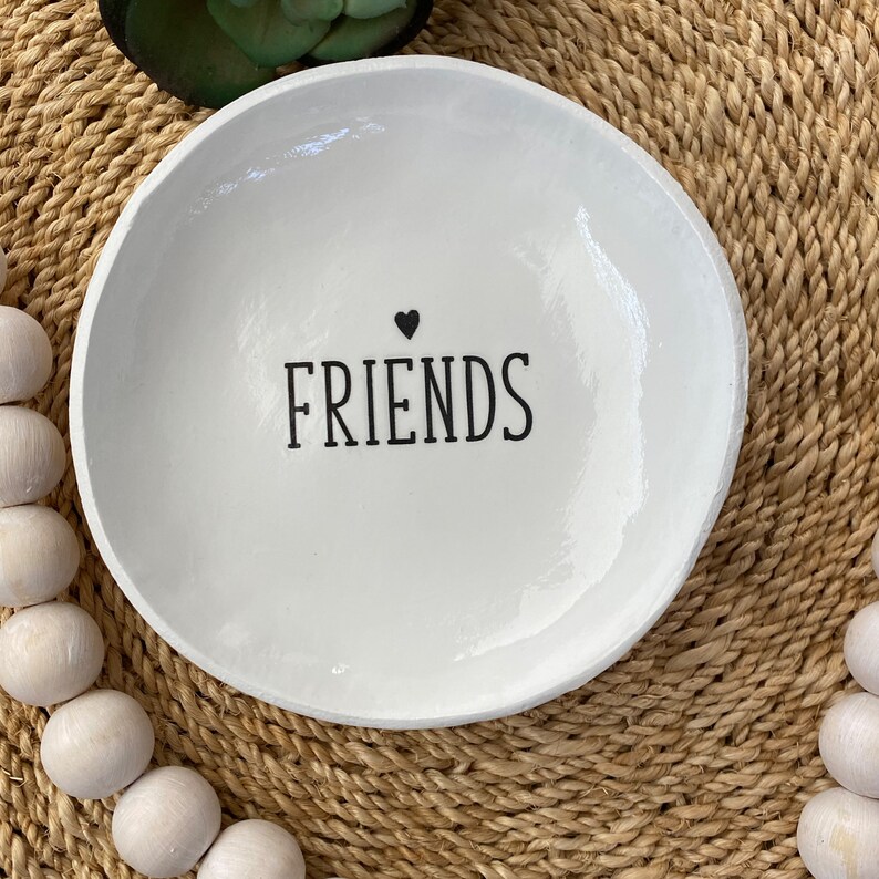 Puede incluir: Un plato decorativo blanco y redondo con la palabra "FRIENDS" en negro y un peque&ntilde;o coraz&oacute;n negro encima. El plato est&aacute; sobre una superficie tejida de color beige, con cuentas de madera blancas a los lados.