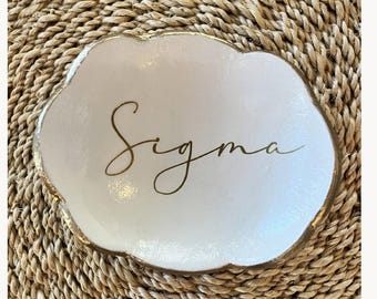Puede incluir: Un plato pequeño blanco con forma de nube y un borde irregular pintado en dorado. El centro del plato presenta la palabra Sigma escrita en una caligrafía dorada. El plato tiene un acabado liso y brillante con un borde festoneado elevado.