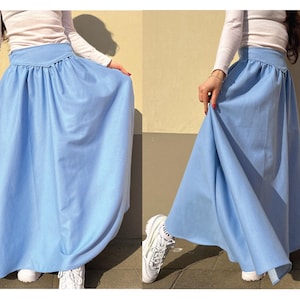 May include: A light blue maxi skirt with a high waist and a flowing silhouette. The skirt is made of a smooth fabric and is paired with a white long-sleeved top and white sneakers. The skirt's design is simple and elegant.