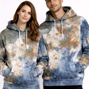 May include: Two people wearing matching tie-dye hoodies. The hoodies feature a pattern of blue, brown, and white hues. Each hoodie has a drawstring hood and a front pocket. The models are standing in front of a white background.