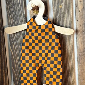 May include: A sleeveless romper featuring a black and orange checkered pattern throughout. The garment has a round neckline and cuffed leg openings that reveal a lighter, muted checkered fabric on the interior lining, displayed on a wooden hanger.