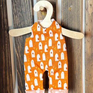 May include: A sleeveless romper featuring an orange fabric with a repeating pattern of white ghosts holding orange pumpkins, small yellow crescent moons, and stars, displayed on a light-colored wooden hanger.