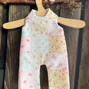 May include: A patchwork baby romper with a floral pattern in pastel pink, yellow, and mint green. The romper is displayed on a wooden hanger against a dark wood background. The fabric is a mix of floral and plaid patterns.