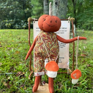 May include: A handmade doll with a pumpkin-shaped head and leaf-patterned overalls. The doll holds a string with two felt mushroom ornaments. A sign behind the doll reads "NEW Z SACKZ" and other text.