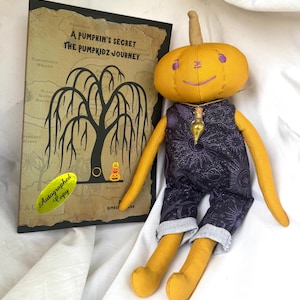 May include: A plush pumpkin doll with a smiling face, wearing a dark purple jumpsuit with a celestial pattern. The doll has long yellow arms and legs. Beside the doll is a book titled "A Pumpkin's Secret The Pumpkidz Journey."
