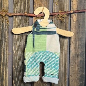 May include: A baby romper with a green, blue, and white geometric pattern hangs on a wooden hanger. The romper is displayed against a rustic wooden background, with a twig and moss accent.