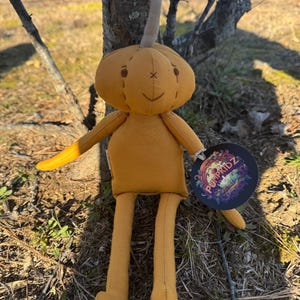 Golden yellow 14-inch Pumpkidz plush doll standing outdoors against a tree, featuring a soft stitched pumpkin face with a calm neutral expression. Handmade adoptable pumpkin plush with fabric tag and Willow’s Cove branding.