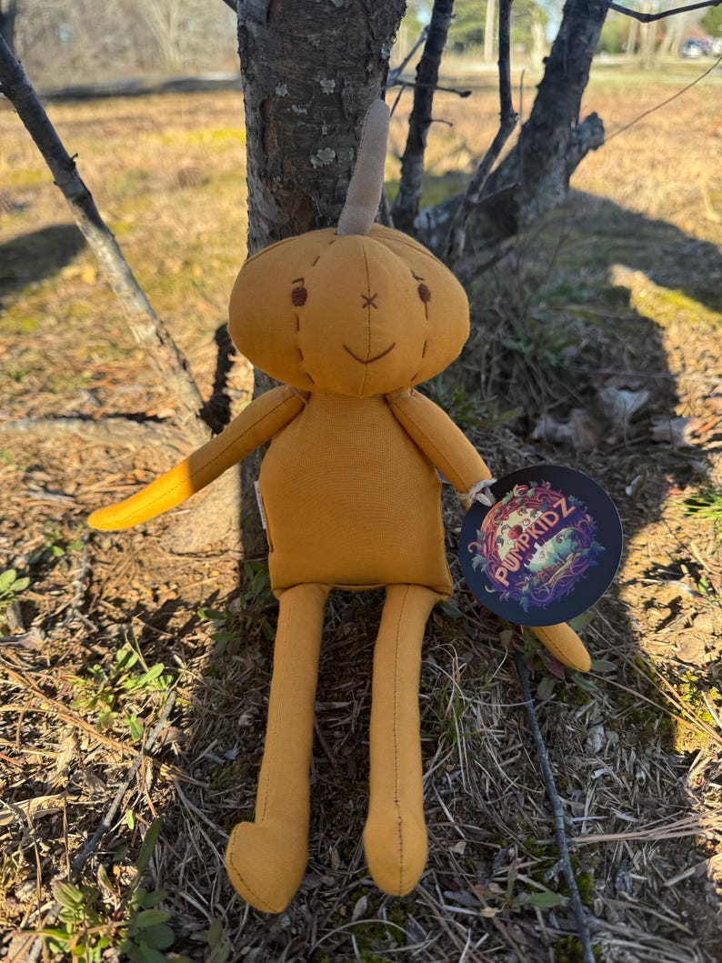 Golden yellow 14-inch Pumpkidz plush doll standing outdoors against a tree, featuring a soft stitched pumpkin face with a calm neutral expression. Handmade adoptable pumpkin plush with fabric tag and Willow’s Cove branding.