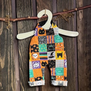 May include: A patchwork Halloween-themed baby romper with orange, black, and white squares. The squares feature Halloween icons like cats, pumpkins, and the words "Trick or Treat." The romper is hanging on a white hanger against a wooden background.