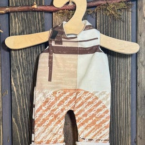 May include: A baby jumpsuit with a geometric pattern in shades of brown, beige, and white, hanging on a wooden hanger. The fabric features a mix of stripes and block designs. The jumpsuit has rolled cuffs at the ankles.