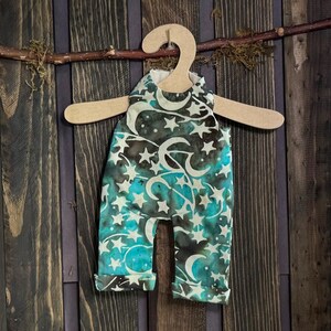 May include: A one-piece baby outfit with a celestial pattern of white stars and crescent moons on a teal and brown background. The outfit is hanging on a wooden hanger against a dark wood background.