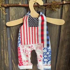 May include: A patriotic baby romper with a patchwork design featuring the American flag, red and white stripes, and floral patterns. The romper is displayed on a wooden hanger against a rustic wooden backdrop.