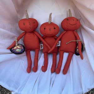 May include: Three red fabric pumpkin-shaped dolls with horns and embroidered faces. The dolls are sitting on a white fabric surface. The dolls are handmade and have a whimsical, folk art style.