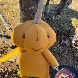 A handmade plush pumpkin toy with a brown stem. The pumpkin is golden yellow and has a stitched-on face with brown eyes, a brown stitched-on smile, and a brown stitched-on X on its forehead. The toy is holding a round tag with the words 'PUMPKIDZ' in a colorful, stylized font.