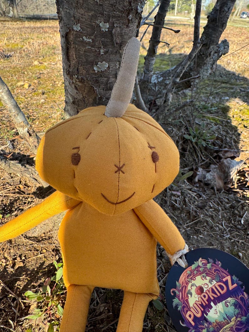A handmade plush pumpkin toy with a brown stem. The pumpkin is golden yellow and has a stitched-on face with brown eyes, a brown stitched-on smile, and a brown stitched-on X on its forehead. The toy is holding a round tag with the words 'PUMPKIDZ' in a colorful, stylized font.