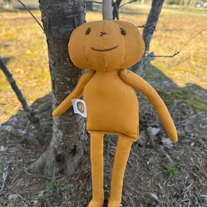 Golden yellow 14-inch Pumpkidz plush doll standing outdoors near a tree, featuring a stitched pumpkin face with a wide gentle smile and cheerful expression. Handmade adoptable pumpkin plush from the Pick-A-Pumpkidz Patch.