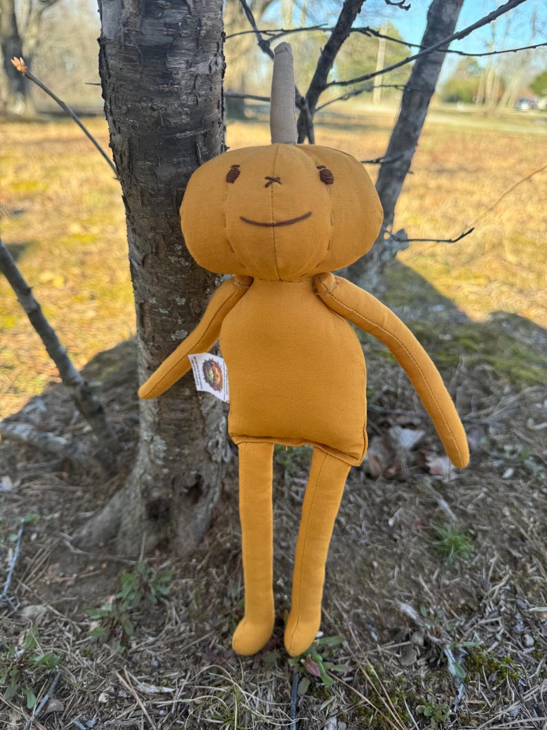 Golden yellow 14-inch Pumpkidz plush doll standing outdoors near a tree, featuring a stitched pumpkin face with a wide gentle smile and cheerful expression. Handmade adoptable pumpkin plush from the Pick-A-Pumpkidz Patch.