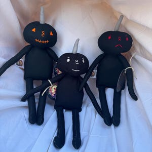 May include: Three black fabric dolls with round heads, thin limbs, and a single gray horn on top of each head. The left doll has orange stitched eyes and a mouth. The center doll has white stitched eyes and a mouth. The right doll has red stitched eyes and a mouth.