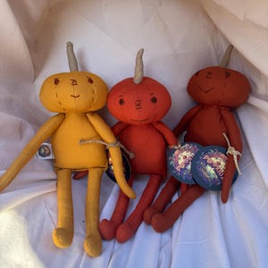 Three handmade pumpkin-shaped plush toys with stitched faces and brown stem. The toys are sitting on a white fabric surface. The toys are in the colors yellow, orange, and dark orange. The toys have tags that say 'Pumpkidz'.
