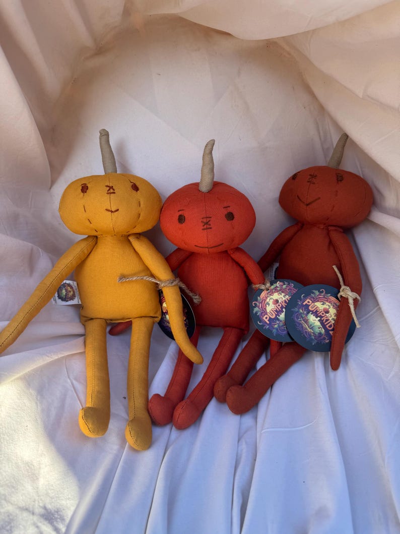 Three handmade pumpkin-shaped plush toys with stitched faces and brown stem. The toys are sitting on a white fabric surface. The toys are in the colors yellow, orange, and dark orange. The toys have tags that say 'Pumpkidz'.